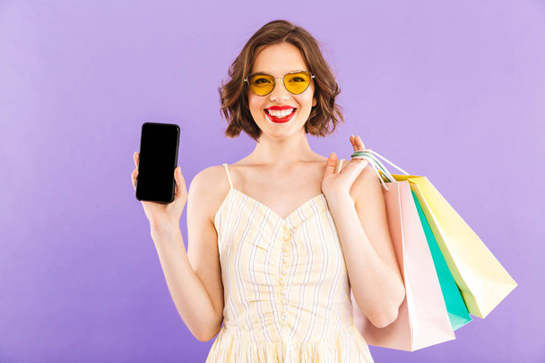 Photo of young woman isolated over purple wall background holding shopping bags showing display of mobile phone. - 写真・画像