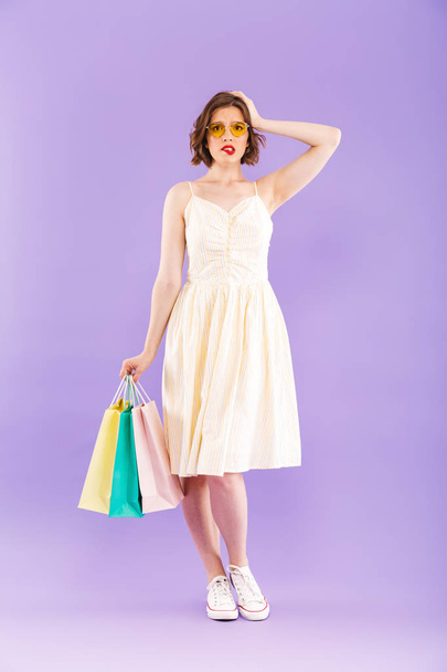 Photo of young confused woman isolated over purple wall background holding shopping bags. - Foto, imagen