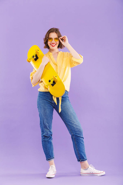 Full length portrait of a cheerful young girl holding skateboard,standing isolated over violet background - Foto, imagen