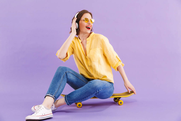 Portrait of a cheerful young girl in headphones sitting on skateboard isolated over violet background - Foto, afbeelding