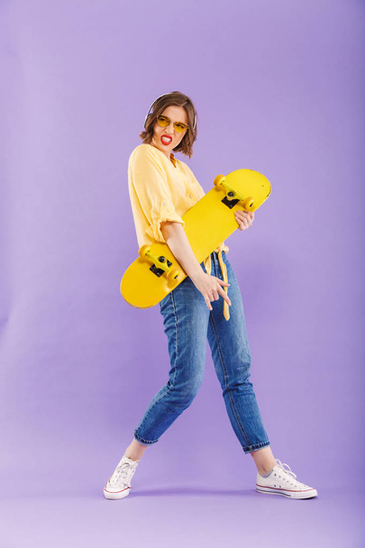 Full length portrait of a cheerful young girl in headphones holding skateboard,standing isolated over violet background - Fotoğraf, Görsel