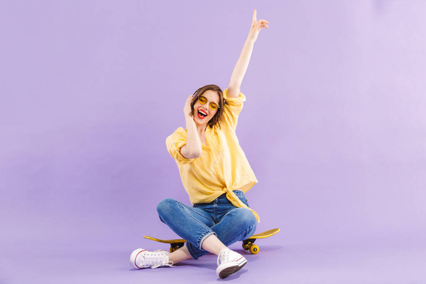 Portrait of a positive young girl in headphones sitting on skateboard isolated over violet background - Фото, изображение