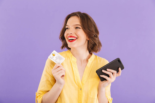 Image of cheerful woman isolated over purple wall background using mobile phone holding credit card. - 写真・画像