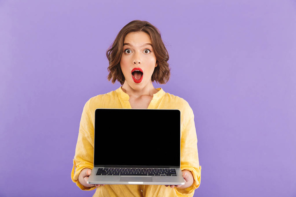 Portrait of a shocked young woman showing blank screen laptop computer isolated over violet background - Foto, imagen
