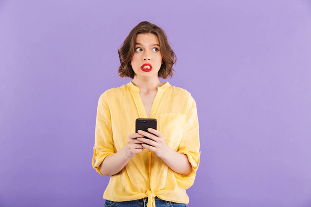 Photo of confused young woman isolated over purple wall background using mobile phone. - Fotoğraf, Görsel