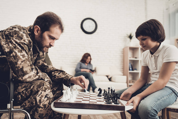 Depressed Paralyzed Veteran Plays Chess With Son. Meeting After War. Wife Background. Camouflage Uniform. Family Concept. Disabled Hero. Home Leisure. Return From Army. Resting Together. - Zdjęcie, obraz