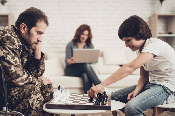 Depressed Paralyzed Veteran Plays Chess With Son. Meeting After War. Wife Background. Camouflage Uniform. Family Concept. Disabled Hero. Home Leisure. Return From Army. Resting Together. - Фото, изображение