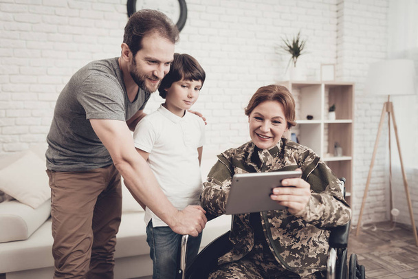Disabled Woman In A Wheelchair Watch A Tablet With Family. Meeting After War. Son And Husband. Camouflage Uniform. Feelings Showing. Return From Army. Paralyzed Soldier. Homecoming Concept. - Foto, Imagem
