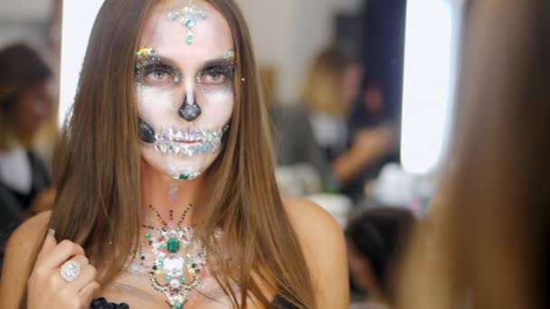Young girl with creative halloween face art looking into mirror reflection in the dressing room. Portrait of glamorous skull with rhinestones and sequins. Professional make-up for the celebration - Footage, Video