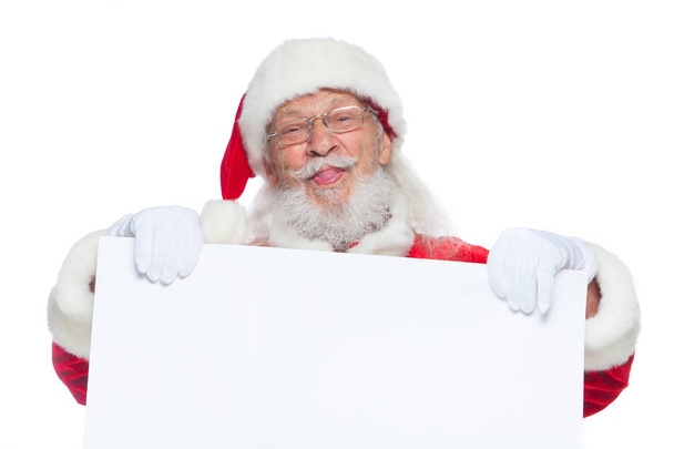 Weihnachten. Der freundliche Weihnachtsmann in weißen Handschuhen mit herausgestreckter Zunge hält einen leeren weißen Karton in der Hand und zeigt Gesichter, Fratzen. Platz für Werbung, für Text, leeren Raum. Copy-Paste. isoliert auf - Foto, Bild