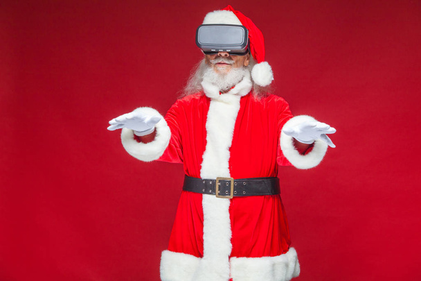 Christmas. Santa Claus in black virtual reality glasses makes gestures with his hands. Surprise, emotion. New technology. Isolated on red background. - 写真・画像