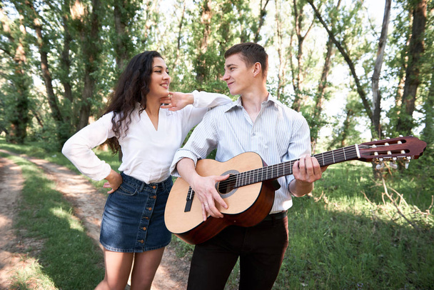 young couple walking in the forest, playing guitar and dancing, summer nature, bright sunlight, shadows and green leaves, romantic feelings - Zdjęcie, obraz