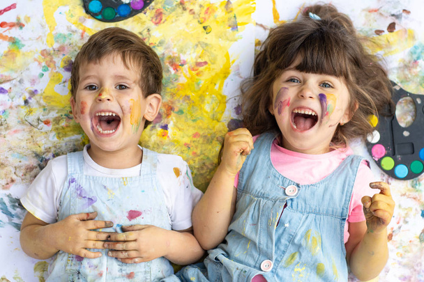 lachende Kinder mit Farbpunkten im Gesicht mit Farben und Pinseln auf dem Farbboden liegend. Kinder malen draußen. - Foto, Bild