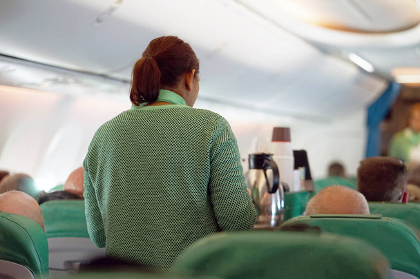 L'hôtesse, au service des passagers, offre thé, café, nourriture pendant le vol. L'intérieur de l'avion avec les passagers assis sur les sièges, et l'hôtesse marchant avec le chariot
. - Photo, image