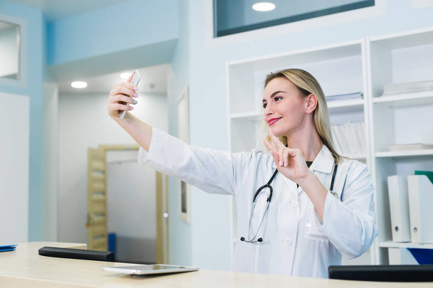 Усміхнена медсестра бере селфі зі своїм телефоном за рецепцією
 - Фото, зображення