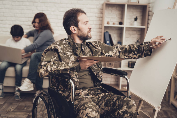 Disabled Soldier In Wheelchair Draws A Picture. After War Concept. Camouflage Uniform. Paralyzed Man With A Tassel. Home Relaxing. Return From Army. Family Background. Wife And Son. - Foto, imagen