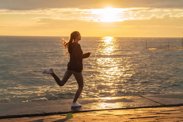 confident slender girl is engaged in jogging at the sea at sunset, preparing for competitions on the run - Фото, изображение
