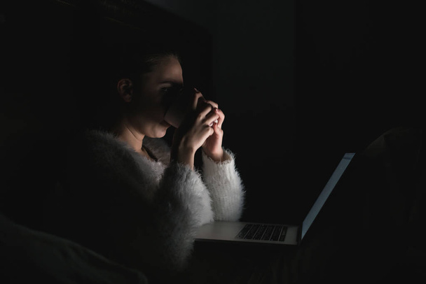 girl freelancer sitting in bed late at night and drinking tea, working on her laptop - Fotoğraf, Görsel