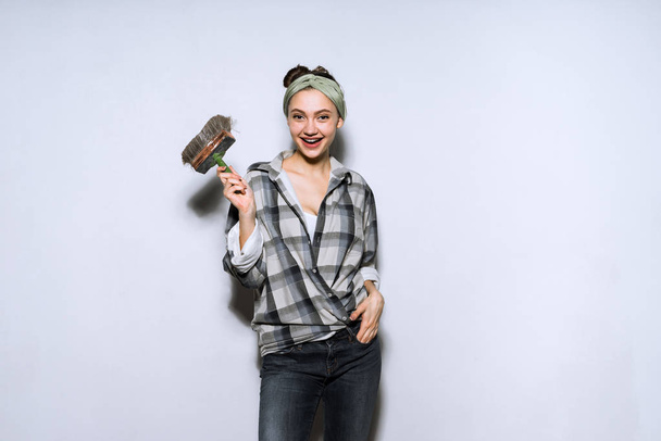 funny young girl in a plaid shirt holding a brush in her hand, doing repairs in an apartment - Foto, afbeelding