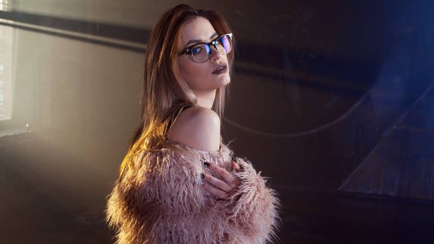 Young seductive woman, portrait in darkness. Girl in stylish pink fur coat and glasses. - Foto, Bild