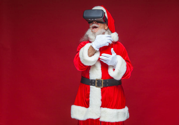 Christmas. Santa Claus in black virtual reality glasses holding a red bucket of popcorn. Watching a movie in virtual reality. Isolated on red background. - Фото, зображення