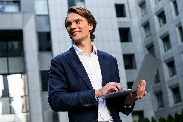 E-Mail unterwegs. schöner junger Mann benutzt seinen Laptop mit einem Lächeln, während er draußen steht - Foto, Bild