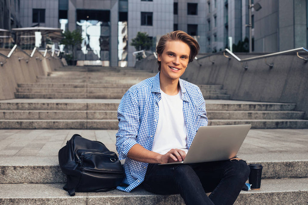Estudiar puede ser divertido! Joven guapo usando su portátil y mirando a la cámara con sonrisa mientras está sentado al aire libre
 - Foto, imagen