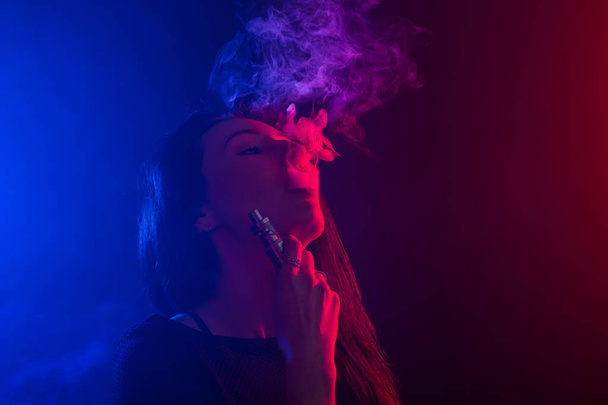 Retrato de mujer asiática fumando vapor o cigarrillo electrónico en luz de neón en fondo negro
. - Foto, Imagen