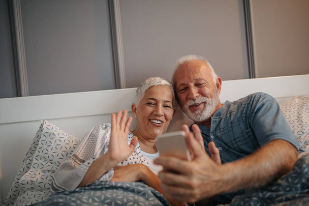 Mature couple lying in bed, holding a mobile phone - Фото, изображение