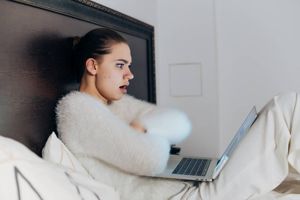 descontenta chica sorprendida en pijama sentada en la cama tarde en la noche y viendo una película en su computadora portátil
 - Foto, Imagen