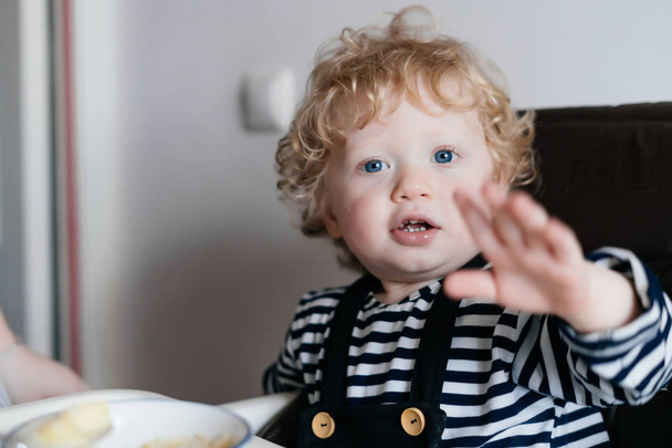 plump little baby boy with blue eyes sitting in the kitchen eating a healthy meal - Foto, Imagem