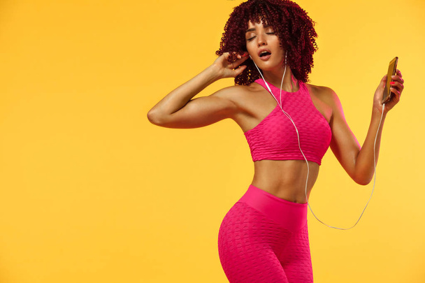 Beautiful young and fit woman in headphones listening music from phone and smiling with closed eyes standing on a yellow background in a pink sportswear - Φωτογραφία, εικόνα