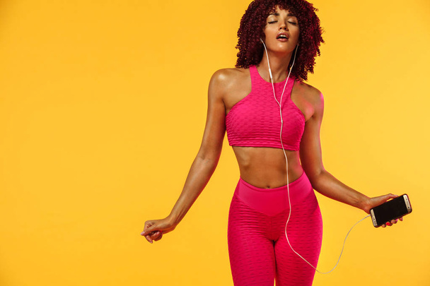 Beautiful young and fit woman in headphones listening music from phone and smiling with closed eyes standing on a yellow background in a pink sportswear - Foto, immagini