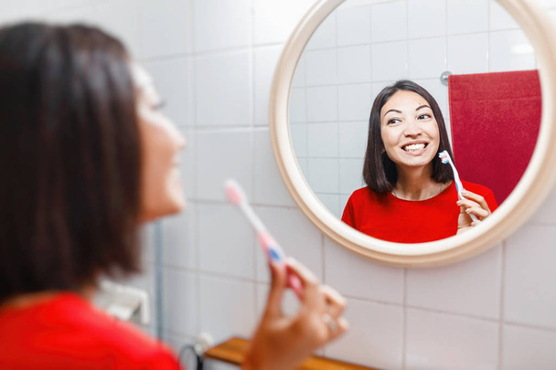Asian woman brushing teeth and looking in the mirror in the bathroom in her apartment - Foto, imagen