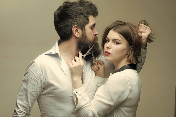 Guy with modern hairstyle visiting hairdresser. Barbershop or hairdresser concept. Man with long beard, mustache and stylish hair, light background. Woman hairdresser cuts beard with scissors. - Фото, зображення