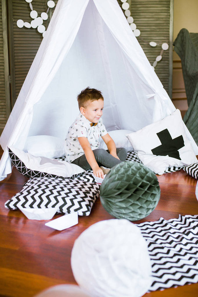 A cute little boy is playing with paper boats at home in a children's tent. Children's room decorated with pompons and paper garlands. Children's holiday - Photo, image
