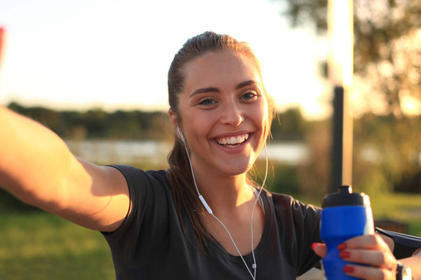 Young attractive woman in sports clothing looking at phone and smiling while taking selfie, during outdoors workout - Fotografie, Obrázek