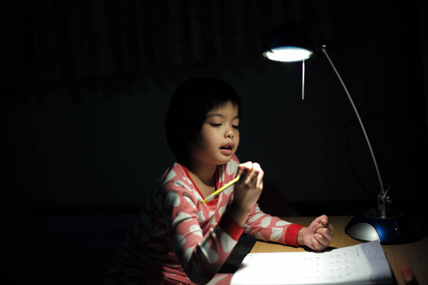 暗い部屋で照明ランプの下で彼女の宿題をしているアジア女の子の肖像画 - 写真・画像