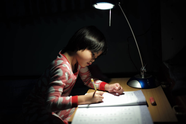 Porträt kleiner asiatischer Mädchen, die ihre Hausaufgaben unter der Lampe in einem dunklen Raum machen - Foto, Bild