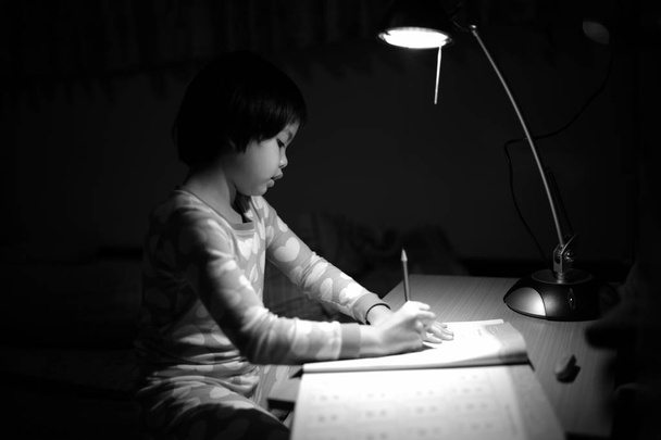 Monochromatyczne portret dziewczynki Azjatki odrabianie pracy domowej w świetle lampy oświetlenie w ciemnym pokoju - Zdjęcie, obraz