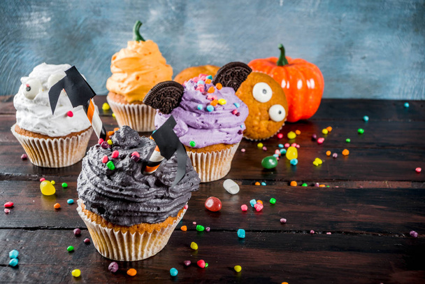 Funny children's treats for Halloween: variations of cupcakes, decorated in the form of different monsters, witches, pumpkins, ghosts. Cooking process. On a wooden table, copy space for text, top view - Photo, Image