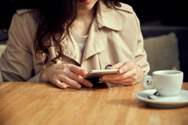 Close up women's hands holding smartphone. Pretty young woman watching video on mobile phone during coffee break, cup of coffee stands near her hands. - Φωτογραφία, εικόνα