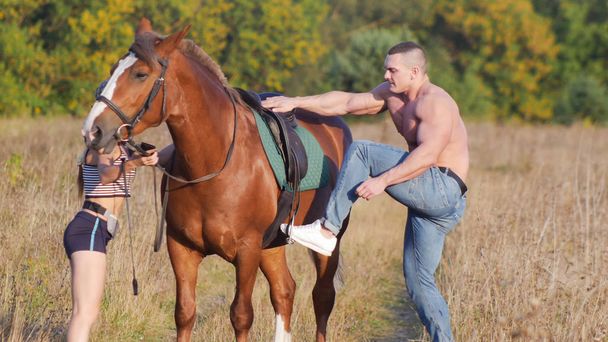 junger Mann mit kräftiger Statur klettert auf ein Pferd, eine junge schlanke Frau hält ein Pferd - Foto, Bild