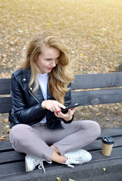 秋の公園で歩いているかなり若い女性リラックス レジャー黒革ジャケット ファッション現代赤い爪飲みコーヒーを離れて - 写真・画像