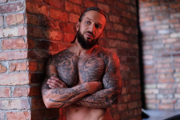 Γενειοφόρος άνδρας με μυϊκή και τατουάζ σώμα ακουμπά σε ένα τοίχο με σταυρωμένα χέρια. - Φωτογραφία, εικόνα