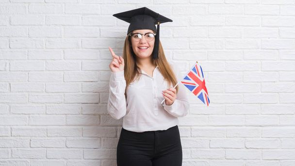 junge erwachsene Frau über der weißen Backsteinmauer mit Absolventenmütze, die die Fahne des Vereinigten Königreichs in der Hand hält, überrascht mit einer Idee oder Frage, die mit glücklichem Gesicht auf den Finger zeigt, Nummer eins - Foto, Bild