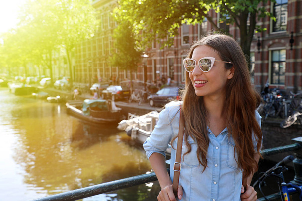 Portrait of beautiful cheerful girl with sunglasses looking to the side on one of typical Amsterdam channels, Netherlands. Light flare vintage filter. - Photo, Image