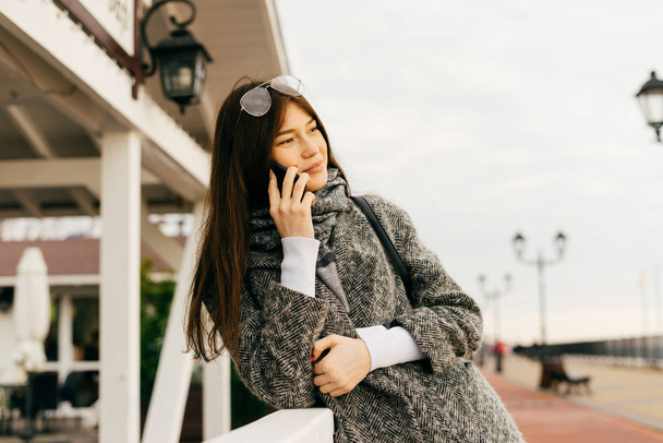 a nice dark-haired girl in a gray coat walking through the streets of the city, enjoying the cloudy weather and talking on the phone - Foto, afbeelding