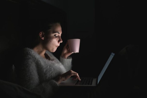 girl freelancer sitting in bed late at night, working on laptop and drinking coffee - Фото, изображение