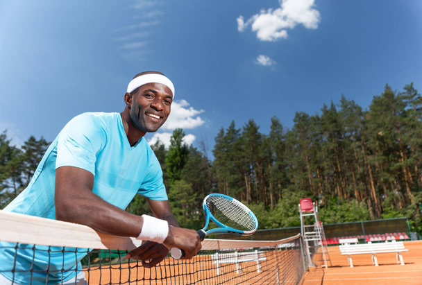 Smiling man is standing and leaning on net. He is holding racket and going to play set. Copy space in right side - Fotografie, Obrázek